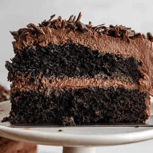 The BEST Fudgy Vegan Chocolate Cake 3 Close-up of a decadent slice of vegan chocolate cake with creamy frosting and chocolate shavings on a white stand.