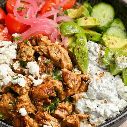 The BEST Chicken Tzatziki Bowl Recipe 2 Vibrant chicken tzatziki bowl featuring seasoned chicken, creamy tzatziki sauce, fresh avocado, cucumber, tomatoes, pickled onions, and crumbled cheese in a dark ceramic bowl.