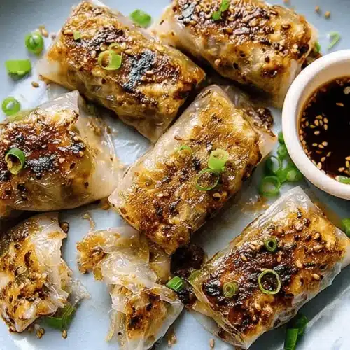 The Best Crispy Rice Paper Dumplings (Pan-Fry or Air-Fry) 2 Golden-brown pan-fried rice paper dumplings with a crispy texture, garnished with sesame seeds and green onions, served with a side of dipping sauce on a light blue plate.