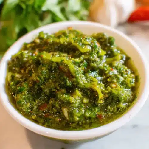 Puerto Rican Sofrito: Authentic Culantro Recipe 2 Close-up of a vibrant green, chunky sofrito in a white bowl, with fresh herbs and peppers blurred in the background, perfect for any sofrito recipe.