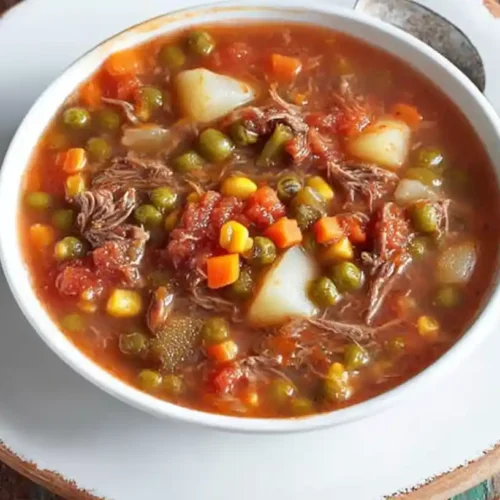 A bowl of vegetable beef soup recipe filled with tender beef chunks, potatoes, peas, corn, and carrots in a rich tomato-based broth, served on a white plate with a spoon beside it.