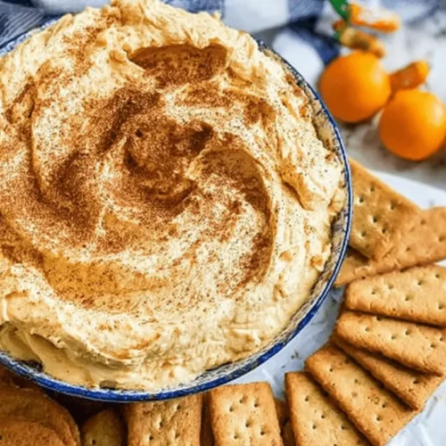 Close-up overhead of creamy pumpkin cheesecake fluff in a white bowl, surrounded by graham crackers, oranges, and yellow spheres on a marble surface.